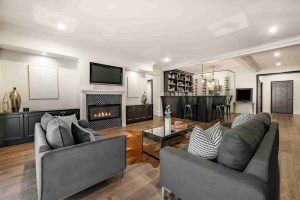 A photo of a luxurious, modern basement living space designed for entertainment. The room features dark wood flooring and a low ceiling with recessed lighting. The sitting area has two dark gray couches facing a fireplace. The fireplace has a black and white patterned tile surround and a mounted TV above it. In the center, a clear glass coffee table rests on a brown and white cowhide rug. In the background, a fully stocked bar area with dark cabinets, open shelving, wine racks, and bar stools is visible.