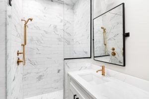 A photo of a luxurious, modern bathroom, featuring white subway tiles with gray marble veining in the shower. The shower is enclosed by a clear glass panel with black framing. Inside the shower, a brass or gold-toned rainfall showerhead and separate handheld shower fixture are mounted on the wall. A white vanity with a matching marble countertop and integrated sink is on the right, with a gold-toned faucet. A rectangular mirror with a thin black frame hangs above the vanity, reflecting the shower.